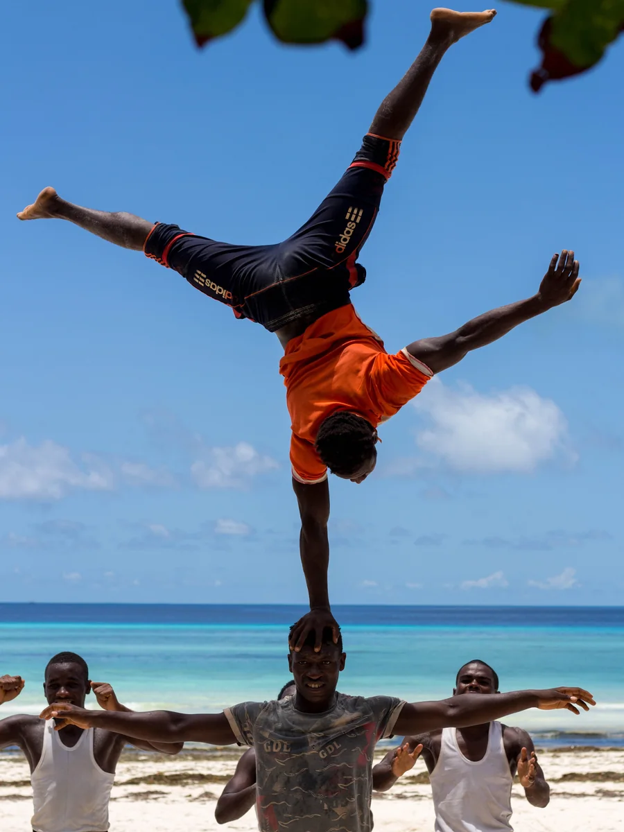 An acrobat performing a one-arm handstand atop a teammate's head, on a beach in Zanzibar, Tanzania (representative photograph of an East African acrobat troupe, not the Safari Cats)